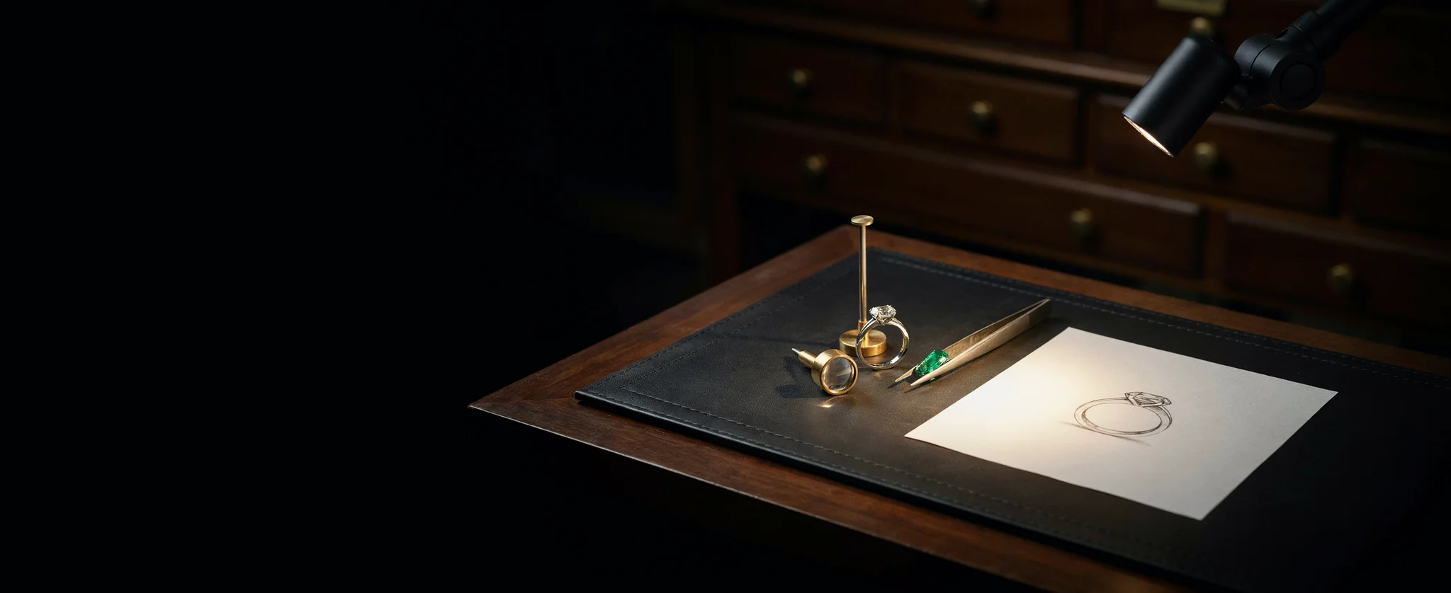 A master jeweler's bench at night: a diamond ring, a loupe, an emerald in tweezers and a pencil drawing of the ring under one lamp (illustrative, generated)